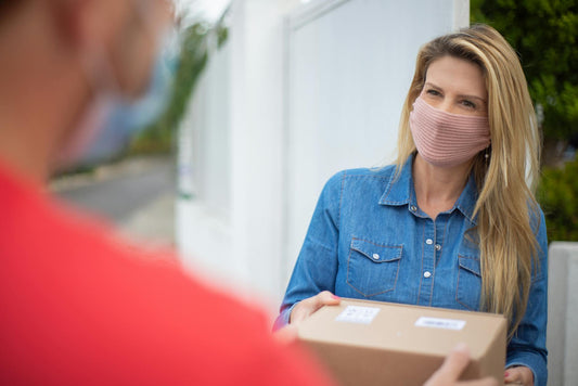 A smiling woman receives a package while wearing a face mask outdoors.