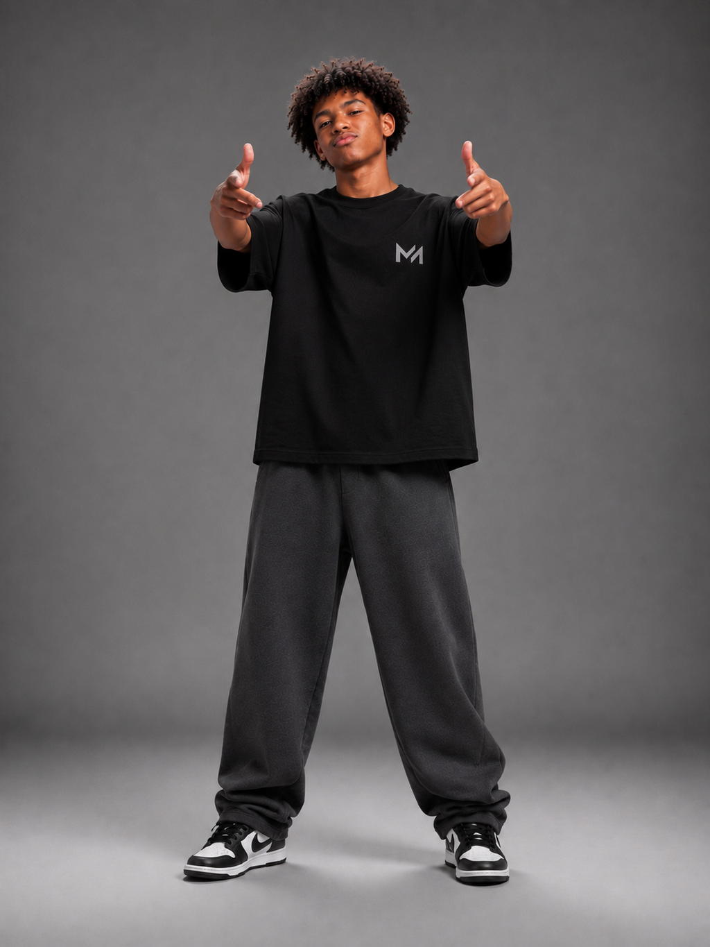 A man modeling a black short-sleeve t-shirt with a small gray 'MILAИI' logo on the left chest, paired with gray sweatpants and sneakers, set against a solid dark gray studio background.