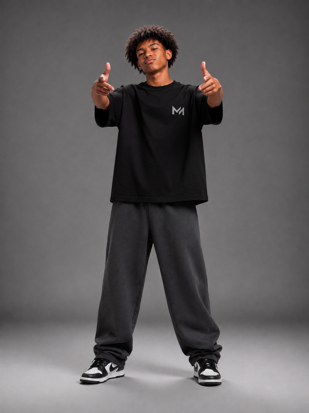 A man modeling a black short-sleeve t-shirt with a small gray 'MILAИI' logo on the left chest, paired with gray sweatpants and sneakers, set against a solid dark gray studio background.