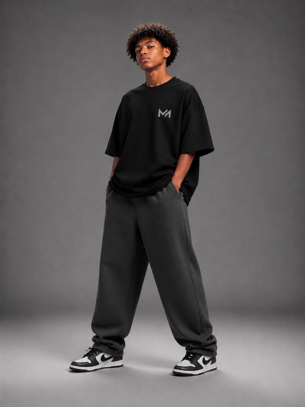 A model stands in a studio wearing an oversized black t-shirt with a small white 'MILAИI' logo on the chest, dark gray sweatpants, and black and white sneakers, against a dark gray background.