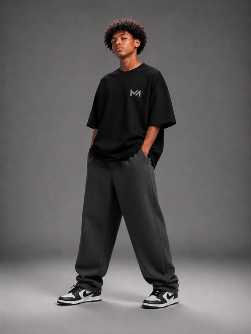 A model stands in a studio wearing an oversized black t-shirt with a small white 'MILAИI' logo on the chest, dark gray sweatpants, and black and white sneakers, against a dark gray background.