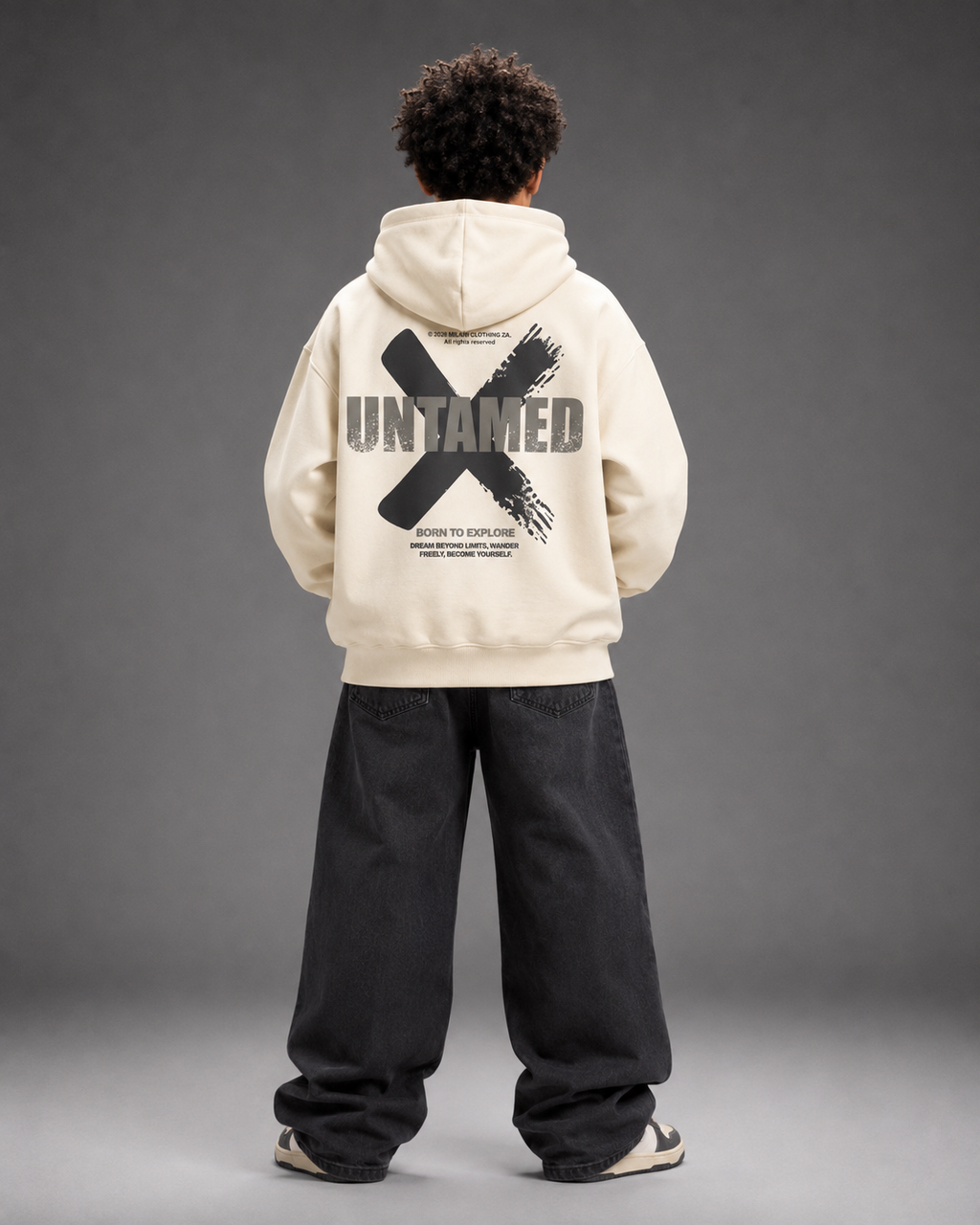 A model viewed from behind wearing a cream-colored hoodie with a large black graphic on the back that reads 'UNTAMED' and 'SHOW TO EXPLORE', paired with loose black pants against a gray studio background.