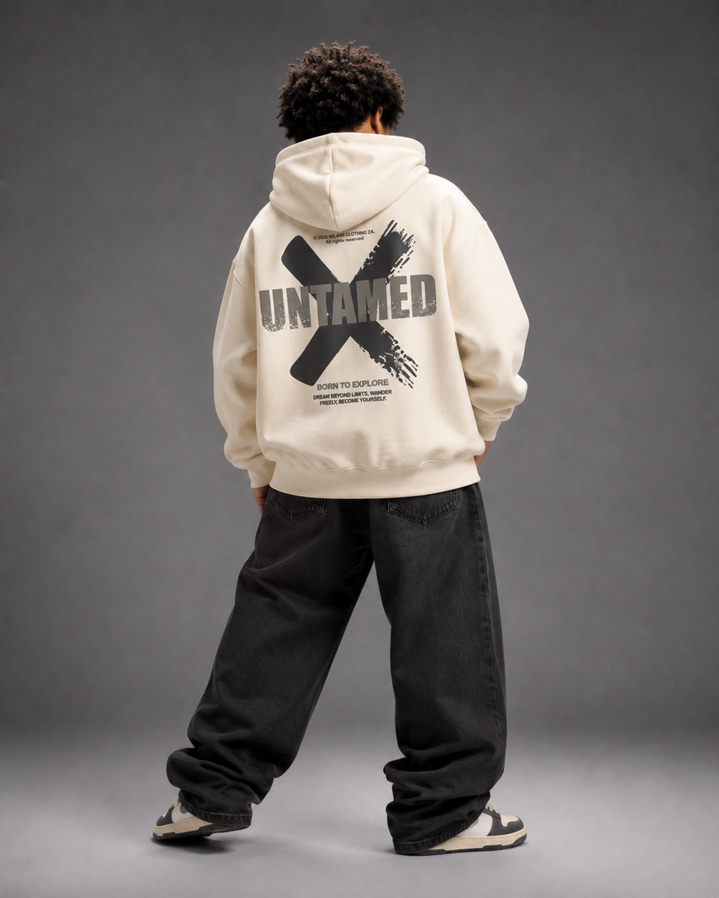A model shown from behind wearing a cream-colored hoodie with a large black graphic print on the back, paired with loose black jeans and sneakers, set against a dark gray studio background.