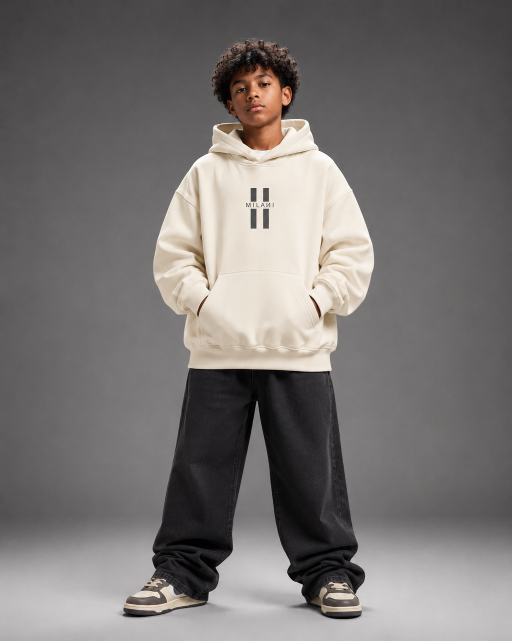 A child model wearing a cream-colored hoodie with a black double-stripe logo on the chest, paired with black sweatpants and sneakers, standing against a dark gray studio background.