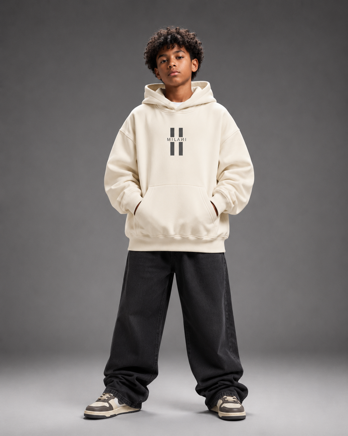 A child model wearing a cream-colored hoodie with a black double-stripe logo on the chest, paired with black sweatpants and sneakers, standing against a dark gray studio background.