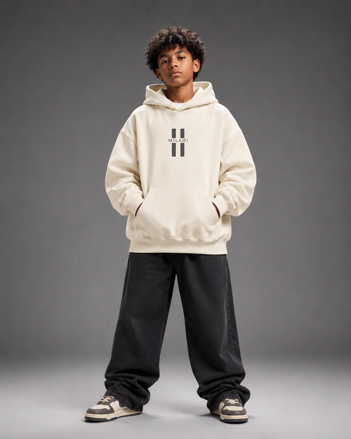 A child model wearing a cream-colored hoodie with a black double-stripe logo on the chest, paired with black sweatpants and sneakers, standing against a dark gray studio background.