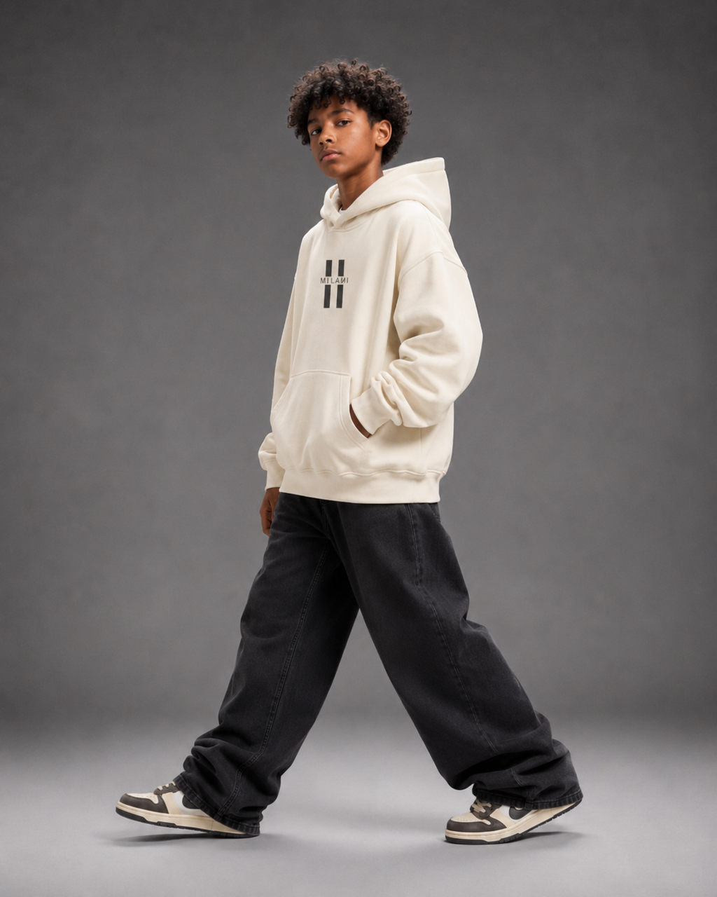 A child model wearing a cream-colored hoodie with a black double-hatch logo on the chest, paired with black baggy sweatpants and sneakers, posing against a dark gray studio background.