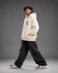 A child model wearing a cream-colored hoodie with a black double-hatch logo on the chest, paired with black baggy sweatpants and sneakers, posing against a dark gray studio background.