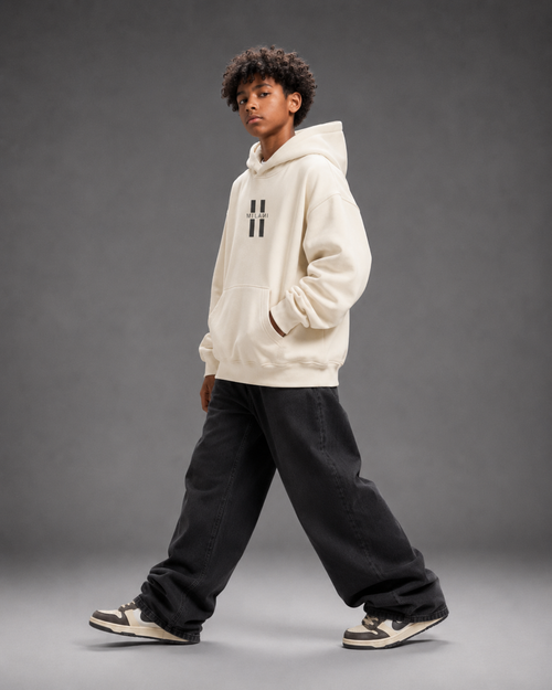 A child model wearing a cream-colored hoodie with a black double-hatch logo on the chest, paired with black baggy sweatpants and sneakers, posing against a dark gray studio background.