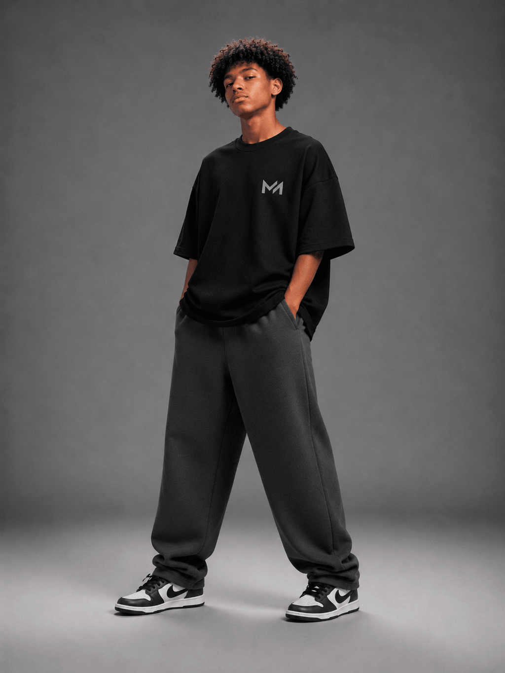 MILANI Born for Stardom Oversized T-Shirt – Black - MILANI CLOTHINGMILANI Born for Stardom Oversized T-Shirt – BlackChildren's Oversized T-ShirtsMILANI CLOTHINGMILANI CLOTHING2-3 yearsA model stands in a studio wearing an oversized black t-shirt with a small white 'MILAИI' logo on the chest, dark gray sweatpants, and black and white sneakers, against a dark gray background.