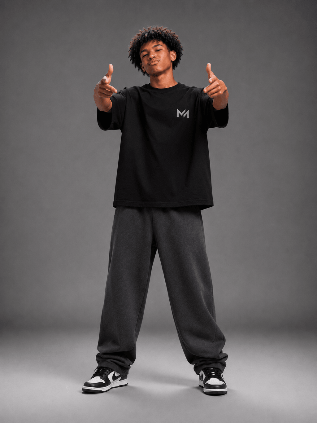 MILANI Born for Stardom Oversized T-Shirt – Black - MILANI CLOTHINGMILANI Born for Stardom Oversized T-Shirt – BlackChildren's Oversized T-ShirtsMILANI CLOTHINGMILANI CLOTHING2-3 yearsA man modeling a black short-sleeve t-shirt with a small gray 'MILAИI' logo on the left chest, paired with gray sweatpants and sneakers, set against a solid dark gray studio background.