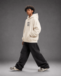 MILANI Untamed Heavyweight Kids Hoodie – Cream - MILANI CLOTHINGMILANI Untamed Heavyweight Kids Hoodie – CreamLoosefit Heavyweight hoodieMILANI CLOTHINGMILANI CLOTHING4-6 YearsA child model wearing a cream-colored hoodie with a black double-hatch logo on the chest, paired with black baggy sweatpants and sneakers, posing against a dark gray studio background.