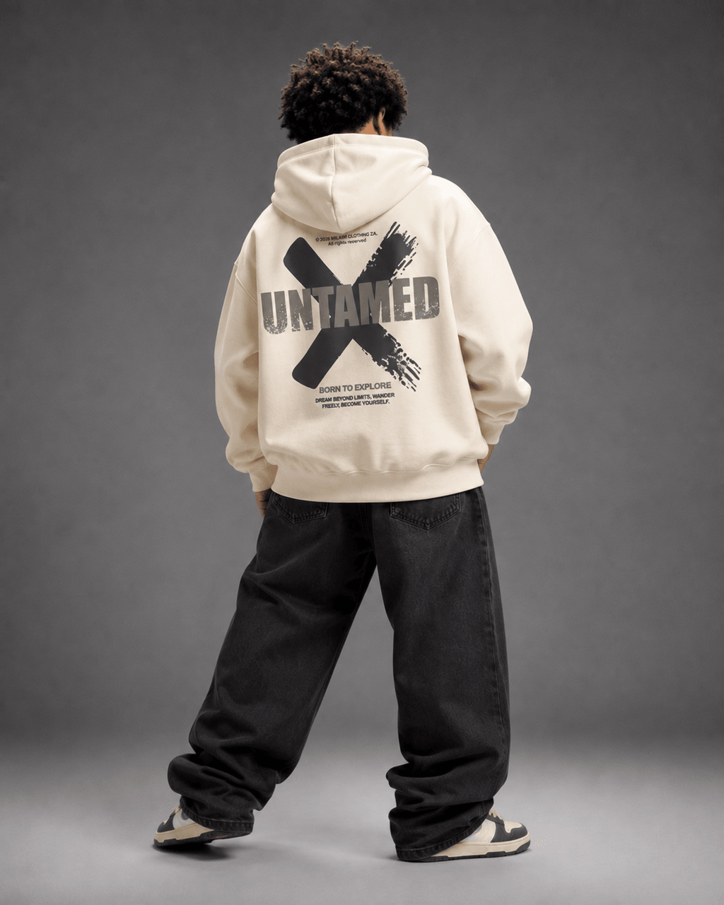 MILANI Untamed Heavyweight Kids Hoodie – Cream - MILANI CLOTHINGMILANI Untamed Heavyweight Kids Hoodie – CreamLoosefit Heavyweight hoodieMILANI CLOTHINGMILANI CLOTHING4-6 YearsA model shown from behind wearing a cream-colored hoodie with a large black graphic print on the back, paired with loose black jeans and sneakers, set against a dark gray studio background.